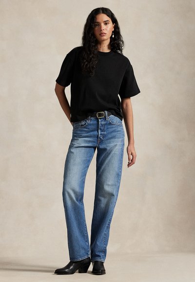 Naine pikkade lokkis juustega, seljas must lühikeste varrukatega särk, sinised teksad ja mustad saapad, seisab neutraalse beezi tausta ees.