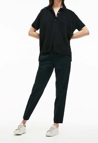 Svart pikétröja med korta ärmar och knappar, kombinerad med mörkgröna avsmalnande byxor. Modellen bär vita sneakers. Enkel och funktionell design.