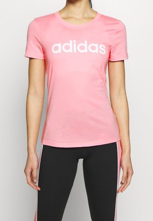 Kobieta ubrana w dopasowany różowy t-shirt Adidas z białym logo oraz czarne legginsy z różowo-białymi paskami po bokach, stojąca na białym tle.