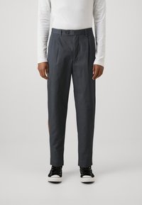 Donkergrijze, op maat gemaakte broek met knoopsluiting, voorplooi, zijzakken en een gestructureerde afwerking. Gecombineerd met zwarte sneakers.