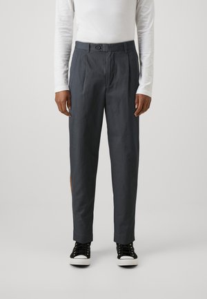 Pantalons ajustés gris foncé avec une fermeture à bouton, des plis à l'avant, des poches latérales et une finition texturée. Associés à des baskets noires.