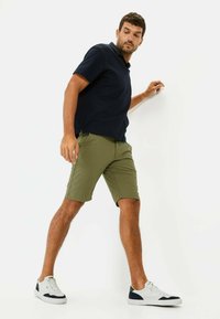 Mannen marineblauwe polo met korte mouwen, gecombineerd met olijfgroene shorts en witte sneakers met marineblauwe accenten. Eenvoudig, casual ontwerp.