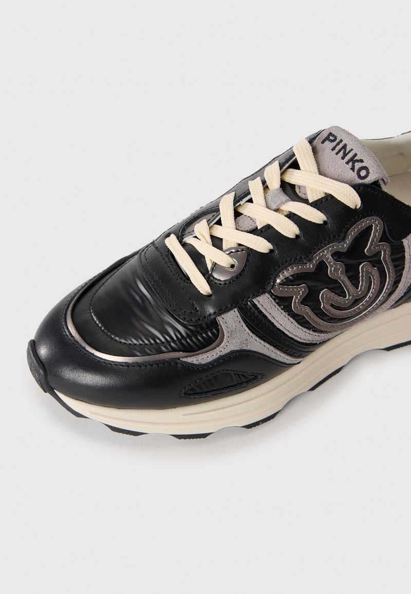 Schwarze Sneaker mit einem glänzenden Lederoberteil, kontrastierenden grauen Akzenten, beigen Schnürsenkeln und dekorativem Logo. Strukturiertes Detail an der Seite.