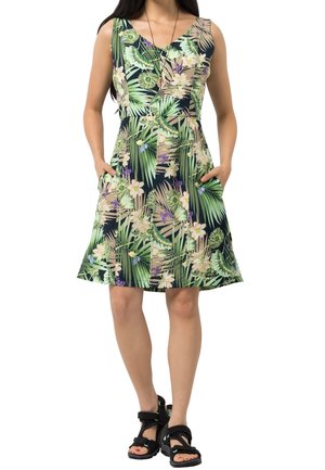 Donna che indossa un abito senza maniche con scollo a V e motivo di foglie e fiori tropicali verdi e beige, mani in tasca e sandali neri con punta aperta.
