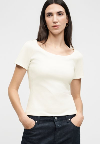 Γυναίκα με μακριά σκούρα μαλλιά, φοράει χρυσούς κρίκους στα αυτιά, ένα στενό λευκό κοντομάνικο μπλουζάκι και σκουρόχρωμο παντελόνι, με τα χέρια στις τσέπες.