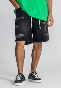 Pantalones cortos de mezclilla negra con detalles desgastados y dobladillo deshilachado. Combinados con una camisa verde y zapatillas negras con cordones blancos.