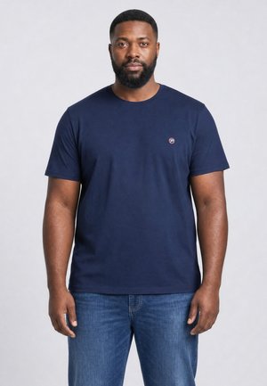 Mand med skæg iført en ensfarvet mørkeblå kortærmet t-shirt og blå jeans står foran en lysegrå baggrund.