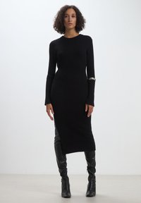 Abito in maglia a coste nera con maniche lunghe e colletto alto, caratterizzato da uno spacco laterale e un dettaglio in argento sull'avambraccio. Silhouette aderente.