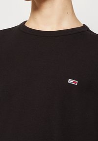 Svart texturerad skjorta med rund hals och en liten broderad logotyp med röda, vita och blå detaljer på vänster bröst.