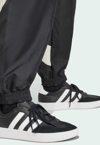 Black athletic pants with elastic cuffs paired with black and white sneakers featuring a textured upper, three white stripes, and white soles.