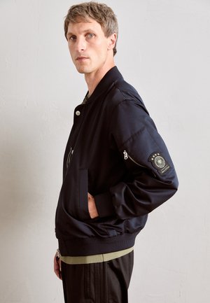 Man wearing a navy bomber jacket with ribbed cuffs and waistband, standing sideways with one hand in pocket against light wall.