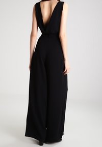 Svart ärmlös jumpsuit med djup V-rygg, vida ben, insvängd midja och en mjuk tygtextur. Enkel och elegant silhuett.