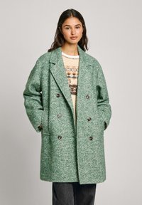 Manteau en motif chevrons vert, croisé avec des boutons bruns, coupe oversize, porté sur un pull crème avec un motif géométrique.