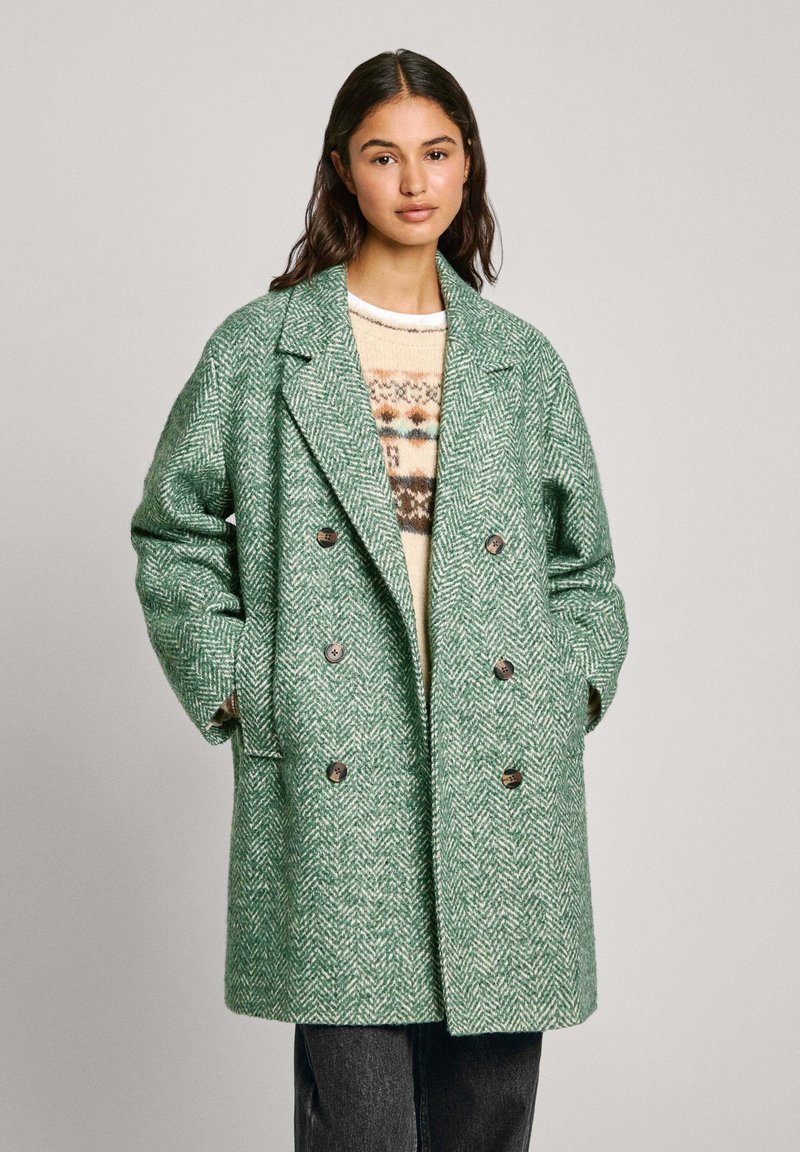 Manteau en motif chevrons vert, croisé avec des boutons bruns, coupe oversize, porté sur un pull crème avec un motif géométrique.