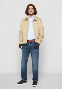 Man with curly hair wearing beige jacket, blue checked shirt, blue jeans, black gloves, brown belt, and navy sneakers standing against white background.