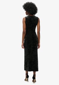 Kobieta z kręconymi włosami, ubrana w bezrękawową czarną welurową sukienkę midi i czarne sandały na obcasie, zwrócona plecami na jednolitym białym tle.