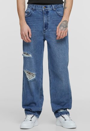 Wide-leg blue denim jeans featuring two large distressed rips, raw hems, and classic front pockets. Paired with white sneakers.