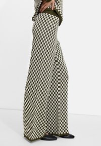 Pantalons à larges jambes à carreaux verts et blancs avec une texture tricotée, dotés d'une taille haute et de poches latérales. Des chaussures noires plates complètent le look.