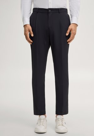 Uomo che indossa pantaloni neri su misura, camicia bianca e sneakers bianche, in piedi davanti a uno sfondo chiaro e semplice.