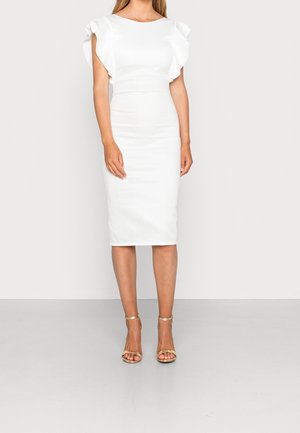 Kobieta w dopasowanej białej sukience do kolan z falbankowymi krótkimi rękawami i złotymi sandałami na wysokim obcasie z paskiem wokół kostki, stojąca na jednolitym tle.