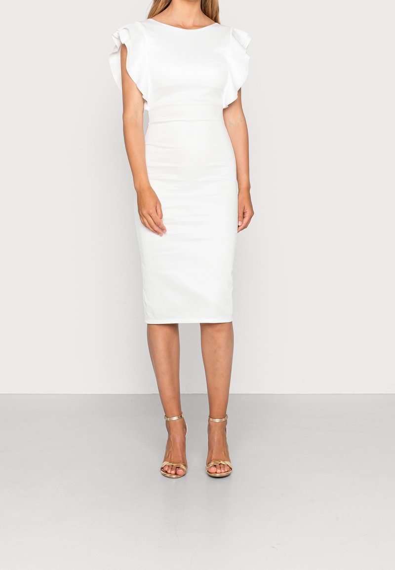 Femme portant une robe blanche ajustée mi-longue aux genoux avec des manches courtes volantées et des sandales à talons hauts dorées avec bride à la cheville, debout sur un fond uni.