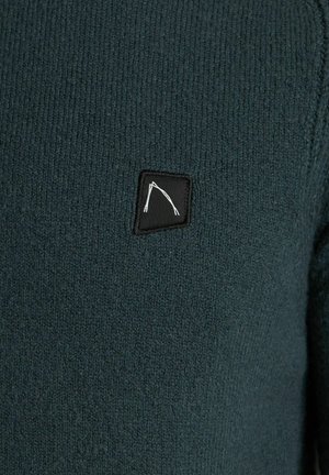 Suéter de punto en verde oscuro con un acabado texturizado y una pequeña etiqueta negra con un diseño gráfico en blanco.