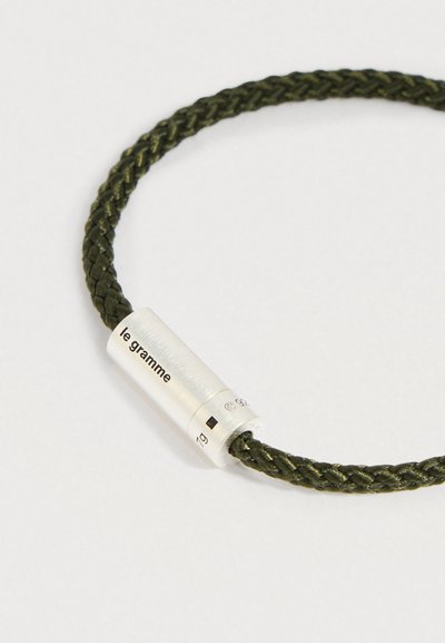 Σκούρο πράσινο πλεκτό βραχιόλι με κυλινδρικό ασημένιο κούμπωμα, χαραγμένο με τη φράση "le gramme" και λεπτομέρειες βάρους, σε λευκό φόντο.