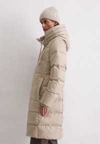 Henkilö, joka käyttää pitkää beigeä untuvakattoa, jossa on korkea kaulus ja huppu, yhdistettynä yhteensopivaan neulottuun pipoon, seisoo sivuttain.