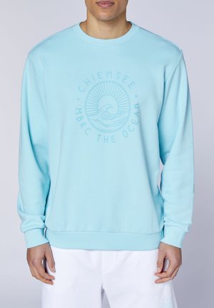 Mann trägt hellblauen Sweatshirt mit Wellen- und Sonnengrafik und Text "Chiemsee MBRC The Ocean", kombiniert mit weißen Hosen.