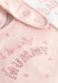 Tessuto rosa chiaro con motivi floreali. Presenta il testo ricamato "Amo la mia MAMMA" in tonalità pastello. Chiusura a scatto sul colletto.