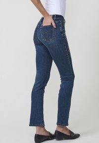 Denim kavbojke v temno modri barvi, z ozkim krojem, vezenimi zadnjimi žepi in rahlimi madeži na robu. Udobno kombinirane z oprijetim zgornjim delom.