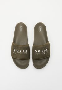 Olivgrüne Slide-Sandalen mit einem strukturierten Netzriemen, der den weißen Text "GUESS EST. 1981" trägt. Glatte Gummisohlen, offenes zehenfreies Design.