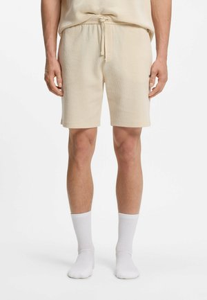 Mężczyzna w beżowych teksturowanych, dzianinowych szortach ze ściągaczem w talii i dopasowanym topem, w połączeniu z białymi skarpetami, na białym tle.