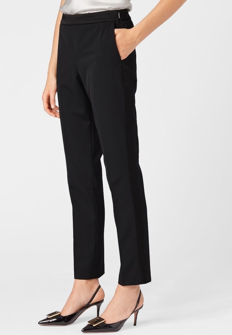 Mustat mukautetut housut, joilla on tiukka muotoilu, sivutaskut ja sileä pinta. Yhdistettynä teräväkärkisiin patentoituihin korkokenkiin, joissa on kultainen yksityiskohta.