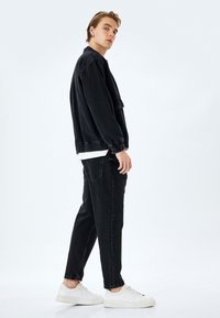 Chaqueta de mezclilla negra con puños elásticos, combinada con jeans negros y zapatillas blancas. El modelo está de lado contra un fondo claro.