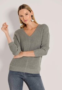 Grauer, gerippter Strickpullover mit V-Ausschnitt und 3/4-Ärmeln. Getragen mit dunklen Jeans, die eine strukturierte Oberfläche und Seitenschlitze aufweisen.