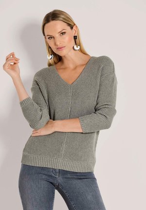 Grauer, gerippter Strickpullover mit V-Ausschnitt und 3/4-Ärmeln. Getragen mit dunklen Jeans, die eine strukturierte Oberfläche und Seitenschlitze aufweisen.