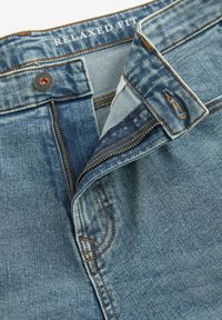Jean de mezclilla azul de corte relajado con costuras visibles, un botón de metal y cremallera. La tela muestra patrones de textura y un diseño clásico.