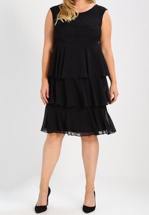 Kobieta w czarnej sukience bez rękawów z falbanami i koronkową górą, w pasujących czarnych szpilkach, stojąca na tle jednolitego jasnego tła.