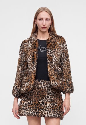 Femme portant une veste en fourrure imprimé léopard et une jupe assortie sur un haut noir avec une écriture en cursive sur un fond clair uni.