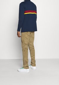 Marinblå långärmad polo med röd och gul horisontell rand, tillsammans med khakifärgade manchesterbyxor och vita sneakers med gröna snören.