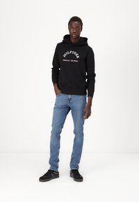 Sort hættejakke med hvid tekstlogo, blå jeans og sorte sneakers. Modellen står foran en ensfarvet baggrund og viser det afslappede outfitdesign.