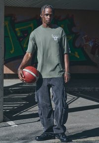 Olivengrøn oversized t-shirt med okse-grafik, parret med sorte cargo-bukser med flere lommer, der holder en rød basketball.
