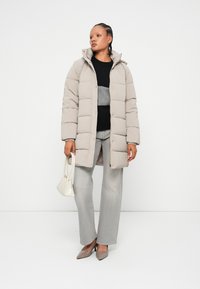 Manteau matelassé beige avec capuche, pull rayé gris, large pantalon gris clair, et escarpins marron clair. Un sac à main blanc complète la tenue.