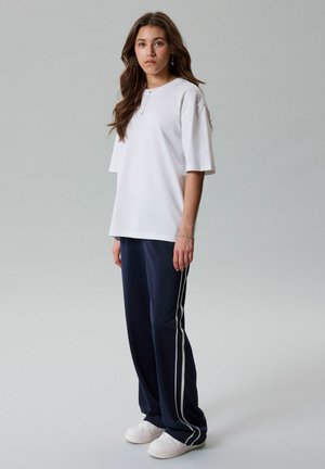 Mladá žena stojí, má na sobě volné bílé tričko, tmavě modré sportovní kalhoty s bílými pruhy a bílé tenisky, na jednobarevném šedém pozadí.