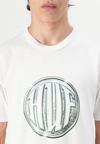 Hvid bomulds t-shirt med en stor cirkulær grafik, der har et "HUF" logo i grøn og grå, sat mod en struktureret baggrund.