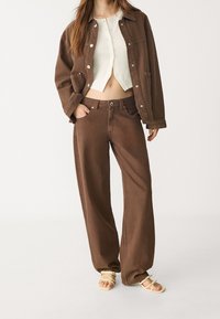 Giacca di denim marrone e pantaloni marroni a gamba larga abbinati, completati da un top corto bianco. Tessuto strutturato con bottoni decorativi. Sandali a completare l'outfit.