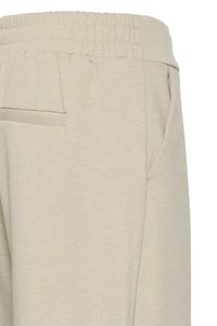 Pantalon beige à taille élastique avec poche latérale sur la couture et détail de poche passepoilée à l'arrière, présenté sur un fond blanc uni.