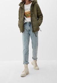Olive green puffer jacket with a hood, white graphic t-shirt, light blue relaxed jeans, and beige lace-up ankle boots with chunky sole.