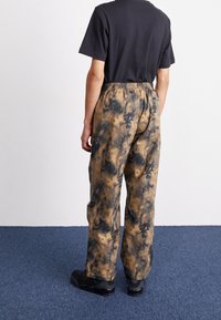 Flerfärgade byxor i en tie-dye-mönster med nyanser av brunt och svart, med en elastisk midja och avslappnad passform. Svarta skor som bärs.